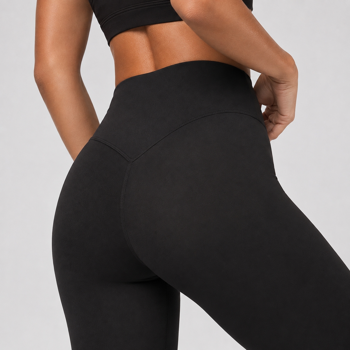 Close-up back view of a model wearing black high-waisted leggings and a matching black sports bra against a light gray studio background.
