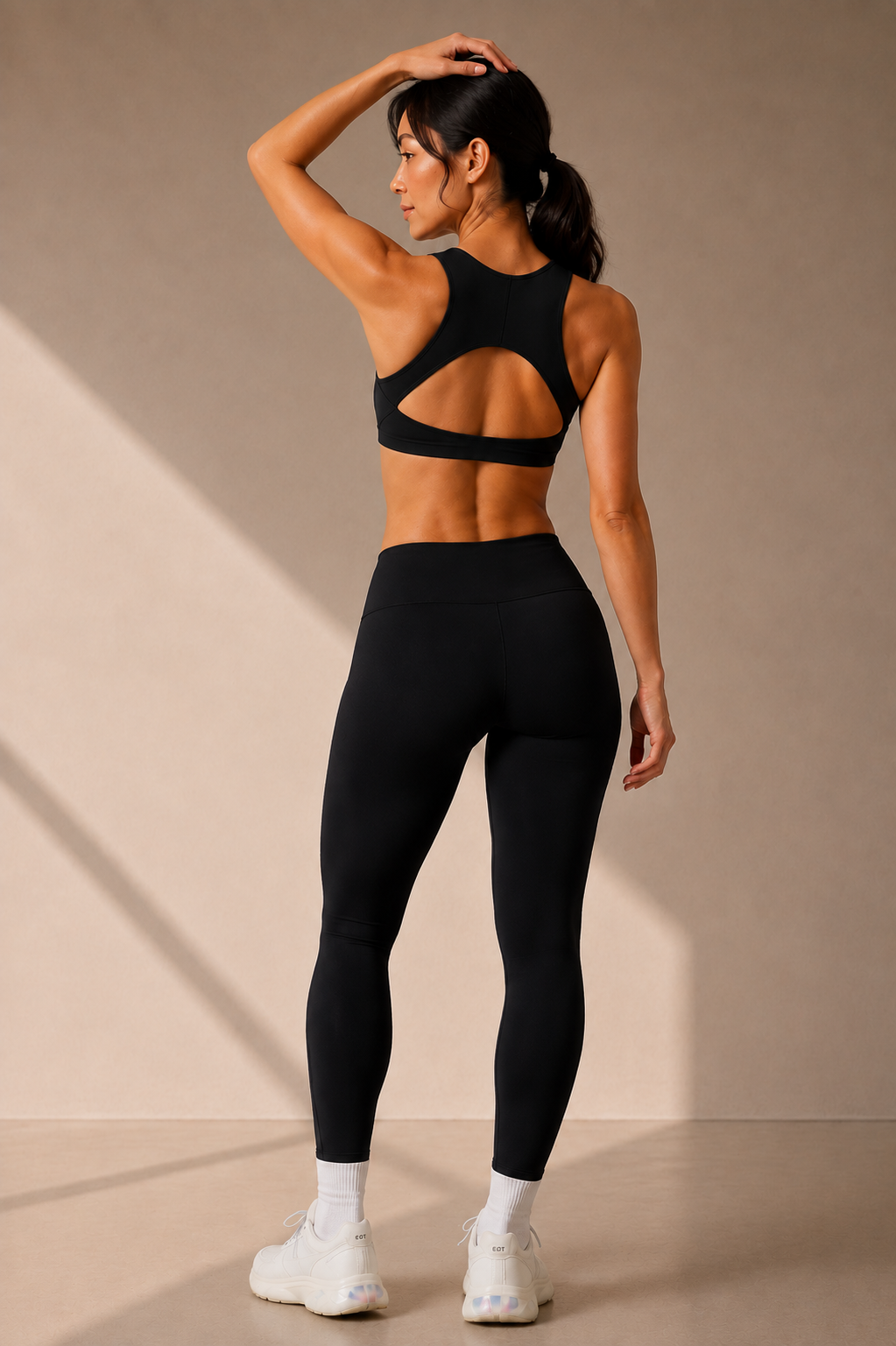 A woman wearing a black sports bra with a large cutout back and matching black high-waisted leggings, shown from the back in a studio setting with a beige wall and floor.