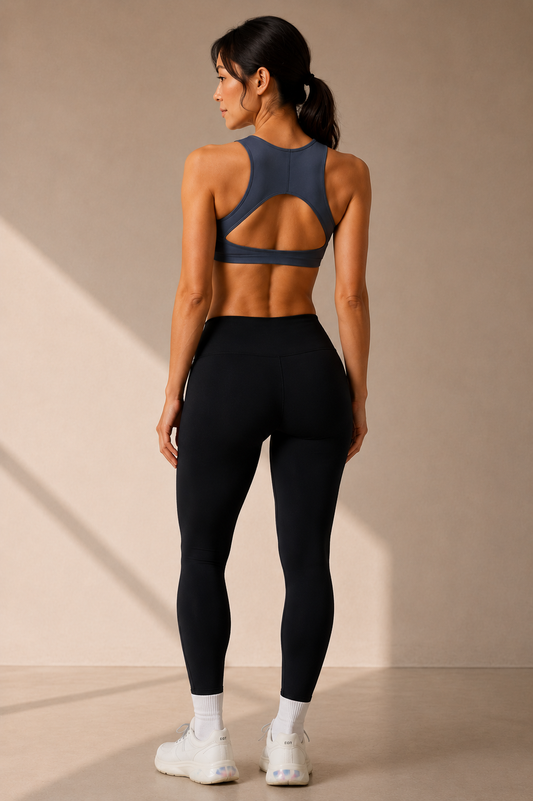 A woman shown from the back wearing a dark slate blue sports bra with a large open-back cutout and black high-waisted leggings, standing in a studio with warm sunlight and shadows.