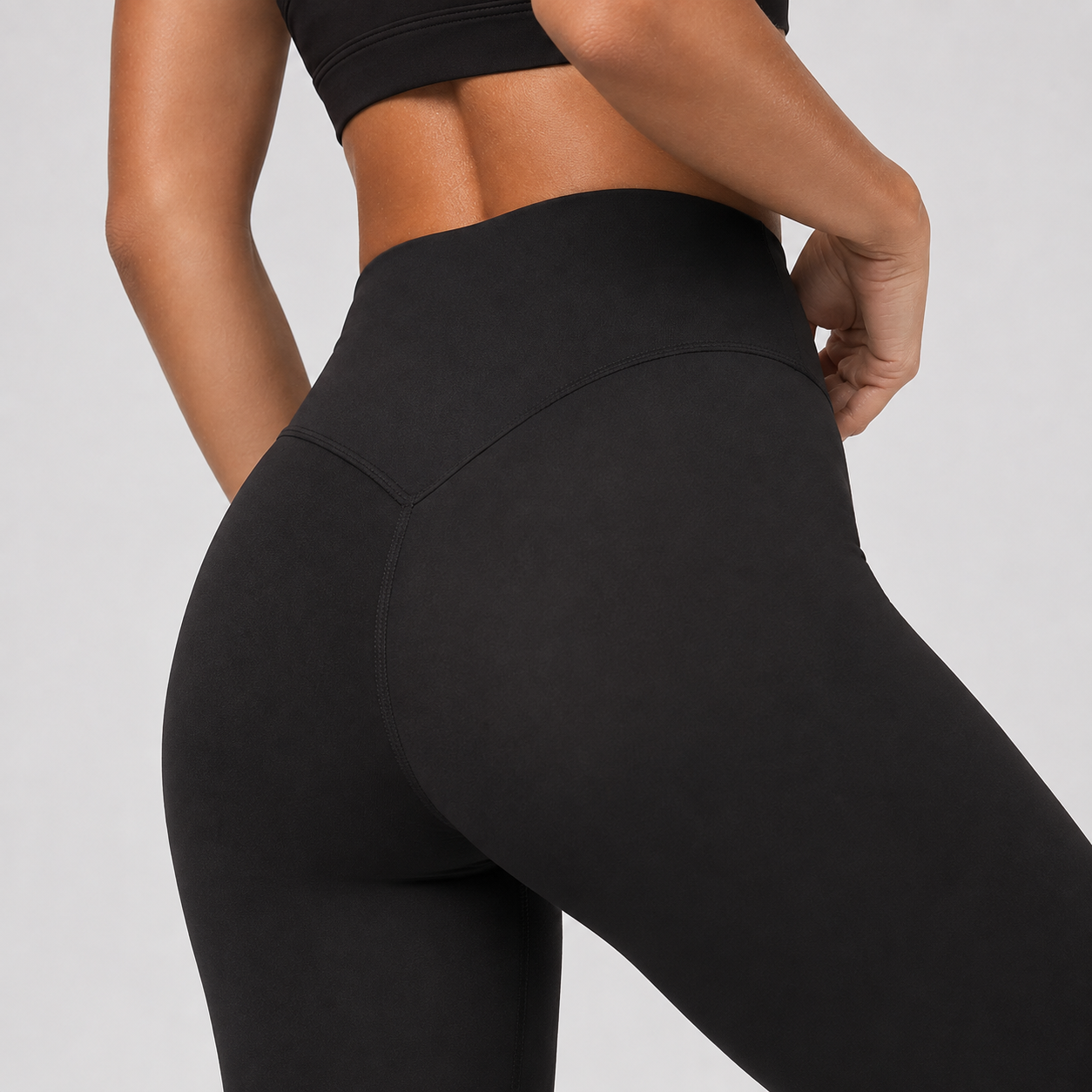Close-up back view of a model wearing black high-waisted leggings and a matching black sports bra against a light gray studio background.