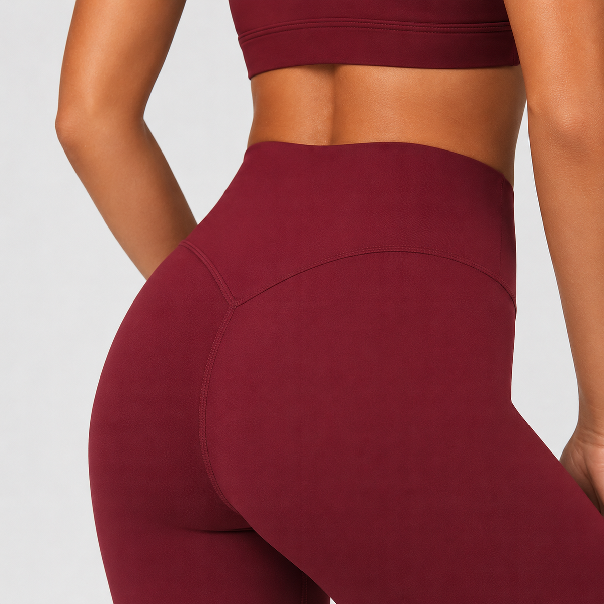 Close-up back view of a model wearing high-waisted burgundy activewear leggings and a matching sports bra against a light gray studio background.
