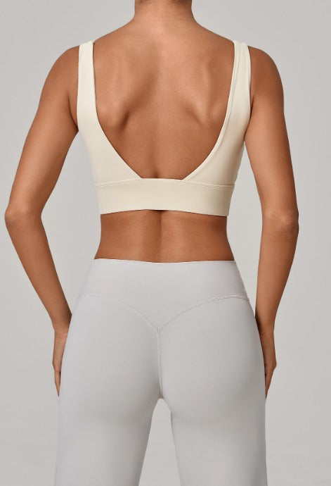 Rear view of a model wearing a light beige sports bra with a wide underband and high-cut light gray leggings against a neutral gray background.